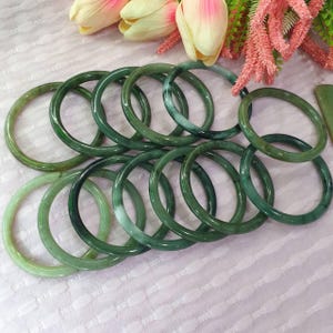 May include: A collection of jade bangles in varying shades of green. The bangles are circular and appear to be made of polished stone. The image is taken from a slightly elevated angle, showcasing the bangles against a soft, light-colored background.