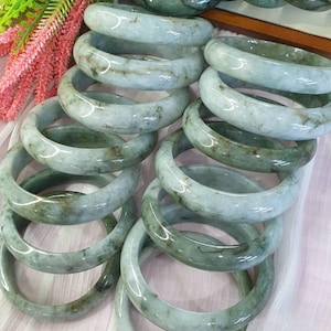 Puede incluir: Una colección de brazaletes de jade en varios tonos de verde y blanco. Los brazaletes pulidos están apilados, mostrando sus patrones naturales y su translucidez. Acentos florales verdes y rosas en el fondo.