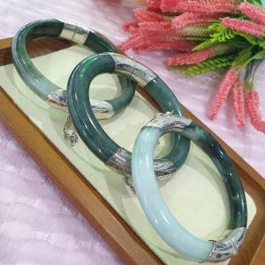 Può includere: Tre braccialetti di giada con dettagli argentati sono esposti su un vassoio di legno. I braccialetti variano nelle tonalità di verde e bianco. Le chiusure argentate presentano disegni intricati. Fiori rosa sullo sfondo.