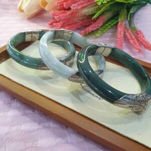 May include: Three jade bangles in varying shades of green and blue-green rest on a wooden tray. Each bangle features silver-colored metal accents. The bangles are displayed on a light-colored surface, with pink and green floral elements in the background.