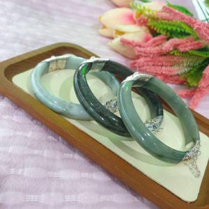 May include: Three jade bangles in shades of green and gray rest on a wooden tray. Each bangle has a silver clasp. The bangles are displayed on a white surface, with pink flowers in the background.