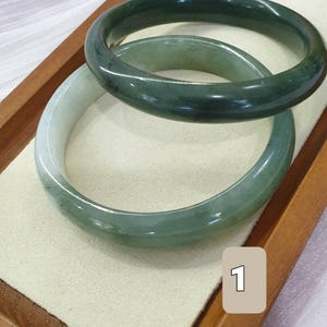 May include: Two jade bangles in shades of green sit on a cream-coloured surface within a wooden tray. The bangles are smooth and polished, with a translucent quality. One is a darker, forest green, and the other is a lighter, more muted green.