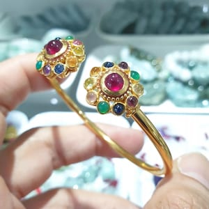 Puede incluir: Pulsera dorada con dos engastes circulares ornamentados. Cada engaste tiene una gema roja central, redonda, rodeada de gemas más pequeñas y coloridas. La pulsera tiene una banda de oro pulido simple.
