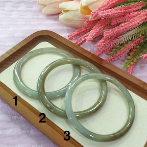 May include: Three jade bangles in varying shades of green and brown are displayed on a wooden tray. The bangles are numbered 1, 2, and 3. The background includes pink and white flowers.