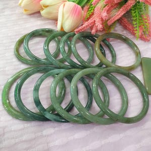 May include: A selection of jade bangles in shades of green. The polished surfaces of the bangles reflect light, highlighting the natural veining of the stone. The bangles are displayed on a pink surface, with floral decorations. The bangles are a popular jewellery item.