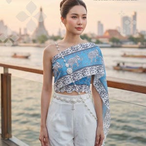 May include: A woman wearing a Thai shawl and outfit. The shawl is light blue with an elephant pattern and white trim. The outfit includes white shorts and a decorative belt. The text "THAI SHAWL" is visible.