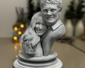 Puede incluir: Una escultura gris de una pareja abrazándose, con rasgos faciales y cabello detallados. La escultura está montada sobre una base redonda y escalonada. El hombre lleva gafas. El tema general es un recuerdo personalizado o un artículo decorativo.