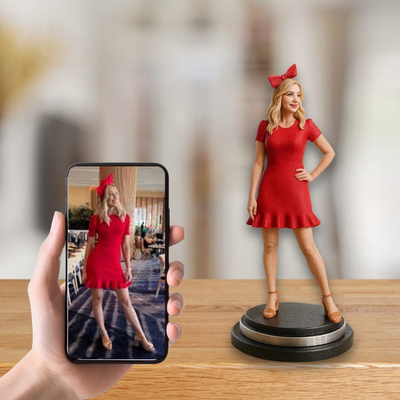 May include: A 3D-printed figurine of a person in a red dress and bow, standing on a black and silver base. The figurine is next to a smartphone displaying a photo of the same person. The dress has short sleeves and a ruffled hem.
