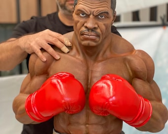 May include: A detailed bust of a muscular boxer with a stern expression, wearing bright red boxing gloves. The sculpture is brown with a textured surface and sits on a gear-like base. The bust is being held by a person.
