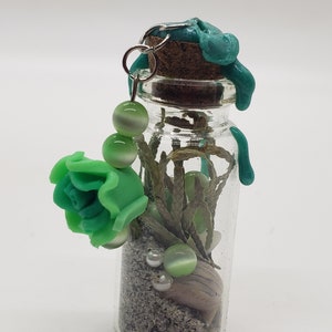 Peut inclure: Une petite bouteille en verre avec un bouchon en liège et un sceau de cire vert. La bouteille contient du sable, une petite coquille, des perles vertes et une fleur en plastique verte.