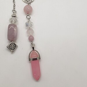 May include: A pink crystal pendulum with a silver chain, a silver pentagram charm, and a silver Celtic knot charm. The pendulum is made of rose quartz and has a pointed tip.