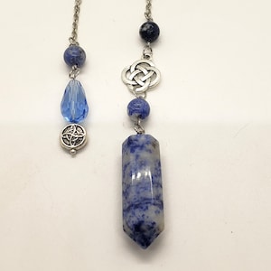 Può includere: Un pendolo di cristallo blu e bianco con una catena d'argento e ciondoli a nodo celtico. Il pendolo è attaccato a una catena d'argento con una perla di cristallo blu e un ciondolo a nodo celtico argentato.