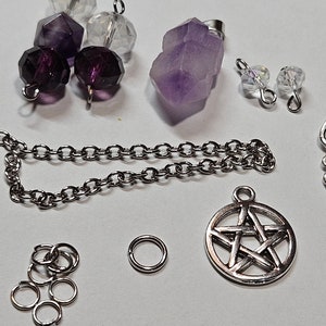 Può includere: Una collezione di forniture per la creazione di gioielli che include una catena d'argento, un ciondolo a forma di simbolo dell'infinito in argento, un ciondolo a forma di pentagramma in argento, un anello di salto in argento, un set di anelli di salto in argento, due perline di cristallo trasparente, tre perline viola e un grande cristallo di ametista viola.