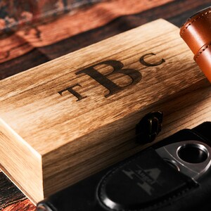 May include: A wooden box with a hinged lid and a brass latch. The box is engraved with the initials "T.B.C.". The box is sitting on a wooden surface next to a black leather cigar case with a silver cigar cutter.