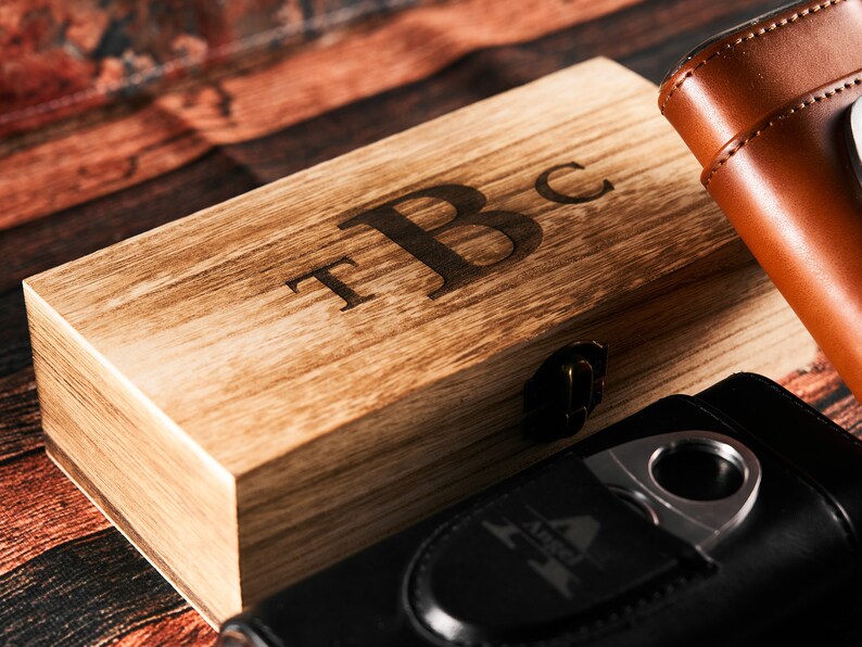 May include: A wooden box with a hinged lid and a brass latch. The box is engraved with the initials "T.B.C.". The box is sitting on a wooden surface next to a black leather cigar case with a silver cigar cutter.