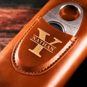 May include: A brown leather cigar case with a built-in cutter. The case has a personalized monogram with the name "Nathan" and the letter "Y".