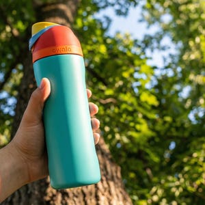 May include: A teal water bottle with a colorful lid, held against a backdrop of green foliage. The lid features yellow, red, and orange sections. The word "awala" is printed on the bottle. The bottle is designed for hydration and outdoor activities.