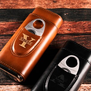 May include: Two leather cigar cases with a built-in cutter. The case on the left is brown with the name "Nathan" and the initial "Y" engraved on it. The case on the right is black with the name "Angel" and the initial "A" engraved on it.