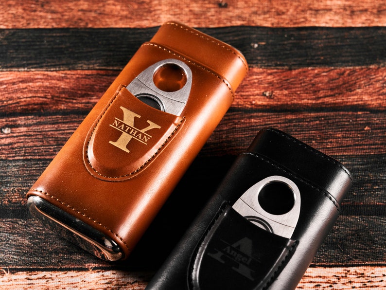 May include: Two leather cigar cases with a built-in cutter. The case on the left is brown with the name "Nathan" and the initial "Y" engraved on it. The case on the right is black with the name "Angel" and the initial "A" engraved on it.