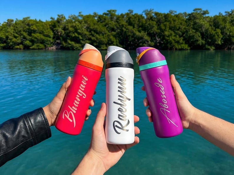 May include: Three insulated water bottles in red, white, and purple are held up against a backdrop of water and trees. Each bottle has a different name printed on it in a cursive font. The bottles have a curved design and a handle on the lid.