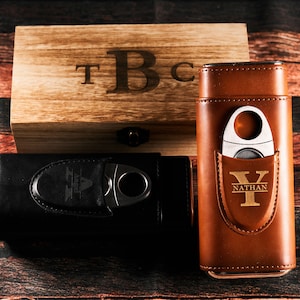 May include: Two personalized cigar cases with a wooden box. One case is black leather with a silver cigar cutter and the initials "A" and "Angel" engraved. The other case is brown leather with a gold cigar cutter and the initials "Y" and "Nathan" engraved. The wooden box has the initials "T", "B", and "C" engraved.