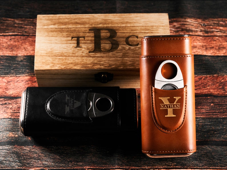 May include: Two personalized cigar cases with a wooden box. One case is black leather with a silver cigar cutter and the initials "A" and "Angel" engraved. The other case is brown leather with a gold cigar cutter and the initials "Y" and "Nathan" engraved. The wooden box has the initials "T", "B", and "C" engraved.
