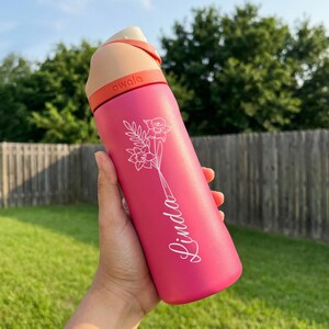 May include: A pink insulated water bottle with a white floral design and the name "Linda" in script. The bottle has a tan lid and an orange carrying loop. The brand name "owala" is printed on the loop.