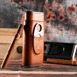 May include: A brown leather cigar case with a built-in cutter and a personalized inscription that reads "Nathan Y". The case is sitting on a wooden surface with a cigar next to it.