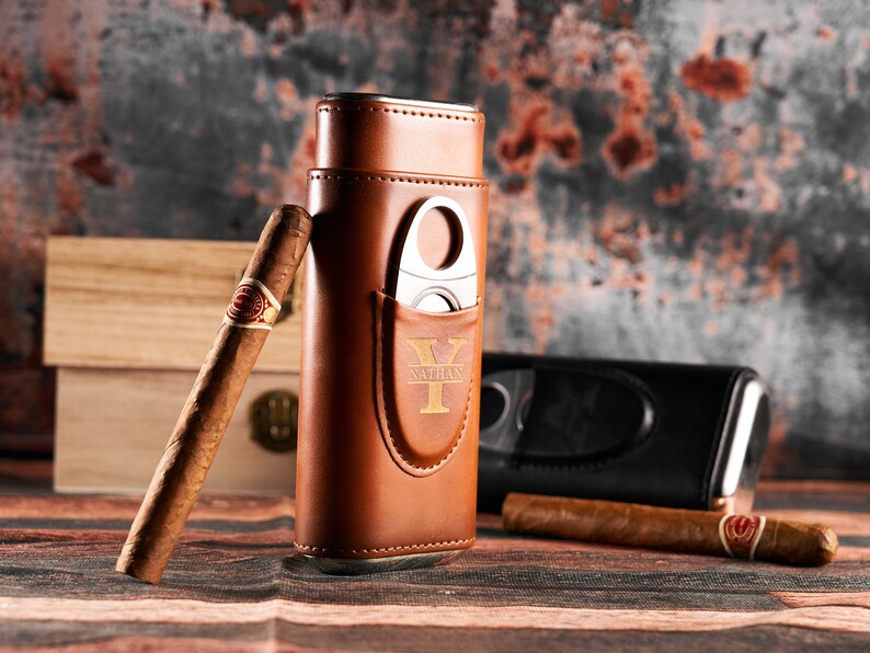 May include: A brown leather cigar case with a built-in cutter and a personalized inscription that reads "Nathan Y". The case is sitting on a wooden surface with a cigar next to it.