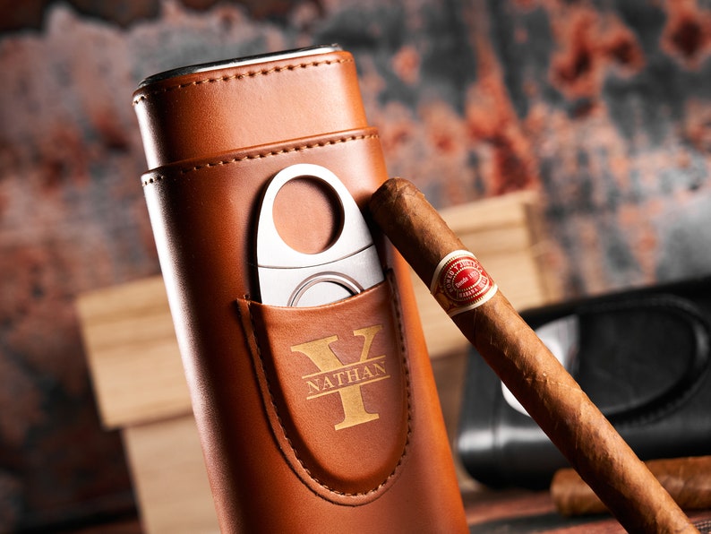 May include: A brown leather cigar case with a built-in cutter and a personalized monogram. The case has a gold Y and the name Nathan engraved on it. A cigar is resting on top of the case.