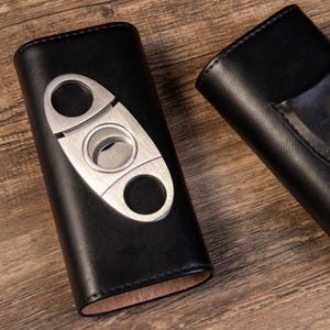 May include: Two black leather cigar cases. One case has a built-in cigar cutter with a silver finish. The other case has a pocket on the outside.