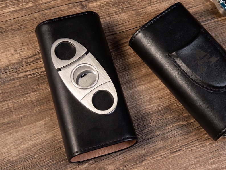 May include: Two black leather cigar cases. One case has a built-in cigar cutter with a silver finish. The other case has a pocket on the outside.