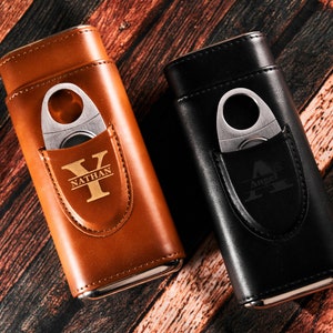 May include: Two personalized leather cigar cases with a built-in cutter. The case on the left is brown with the name "Nathan" and the initial "Y" engraved on it. The case on the right is black with the name "Angel" and the initial "A" engraved on it.