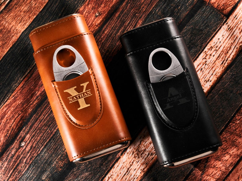 May include: Two personalized leather cigar cases with a built-in cutter. The case on the left is brown with the name "Nathan" and the initial "Y" engraved on it. The case on the right is black with the name "Angel" and the initial "A" engraved on it.