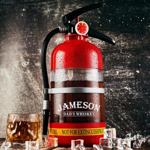 May include: A novelty fire extinguisher-shaped whiskey decanter with the text "JAMESON DAD'S WHISKEY". The red decanter has a black handle and nozzle, with a clear section to view the whiskey. A glass of whiskey and ice cubes are in the foreground.