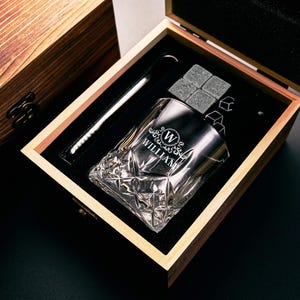 May include: A wooden gift box containing a personalised whiskey glass, ice cubes, and a stirring spoon. The glass is engraved with the name "WILLIAM" and a decorative emblem. The box is open, revealing the contents on a black velvet lining.