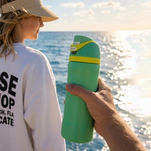 May include: A green water bottle with a yellow accent is held in front of a person on a boat. The person is wearing a white sweatshirt with black text that reads "Sunrise Surf Shop Jacksonville Beach, FLA Surf Syndicate". The background is the ocean.