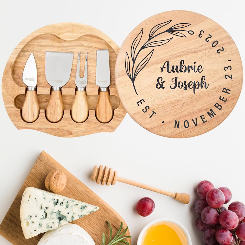 Pode incluir: Uma t&aacute;bua de queijo de madeira com uma parte superior redonda e um design floral. A t&aacute;bua tem o texto "Aubrie & Joseph Est. Novembro 22, 2023" inscrito nela. A t&aacute;bua vem com quatro facas de queijo.
