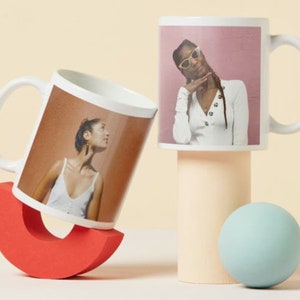 May include: Two white ceramic mugs with handles. The mug on the left has a photo of a person with brown hair and a white tank top. The mug on the right has a photo of a person with brown hair wearing sunglasses and a white shirt. The mugs are on a light beige surface.