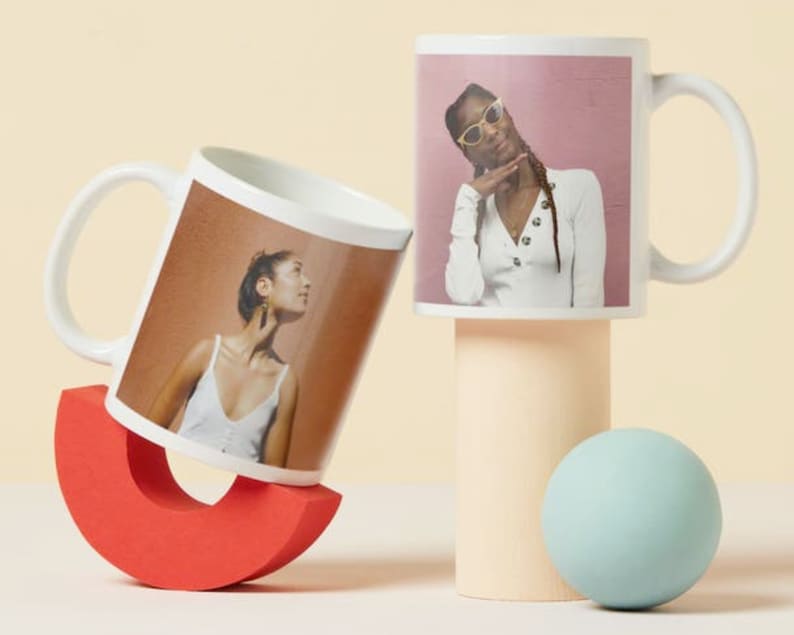 May include: Two white ceramic mugs with handles. The mug on the left has a photo of a person with brown hair and a white tank top. The mug on the right has a photo of a person with brown hair wearing sunglasses and a white shirt. The mugs are on a light beige surface.