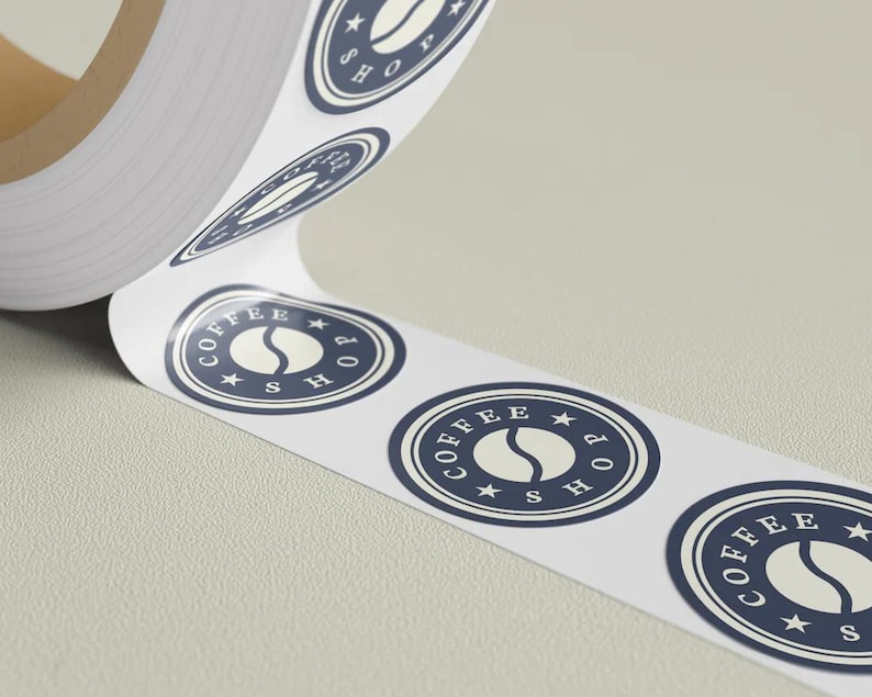 May include: A roll of white stickers with a navy blue and white coffee shop logo. The logo features a coffee bean and the words "COFFEE SHOP" in a circular design. The stickers are on a roll, ready for use.