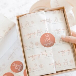 Pu&ograve; includere: Una scatola di cartone marrone con un rivestimento in carta velina bianca. La carta velina ha un motivo ripetuto di un'impronta di zampa di cane e il testo "wagging tails clothing and accessories". Un adesivo circolare rosa con una silhouette di cane e il testo "wagging tails" &egrave; sulla carta velina.
