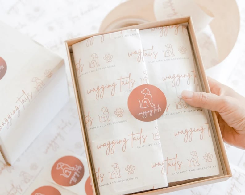 Pu&ograve; includere: Una scatola di cartone marrone con un rivestimento in carta velina bianca. La carta velina ha un motivo ripetuto di un'impronta di zampa di cane e il testo "wagging tails clothing and accessories". Un adesivo circolare rosa con una silhouette di cane e il testo "wagging tails" &egrave; sulla carta velina.