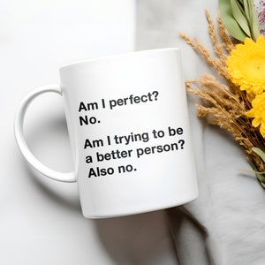May include: White ceramic mug with black text: "Am I perfect? No. Am I trying to be a better person? Also no." The mug is on a light grey surface with yellow flowers and dried plants.