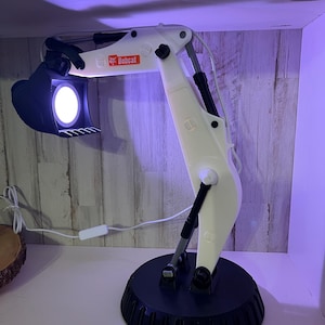 May include: White Bobcat excavator desk lamp with a black base and a white cord. The lamp is turned on and the light is shining from the bucket.