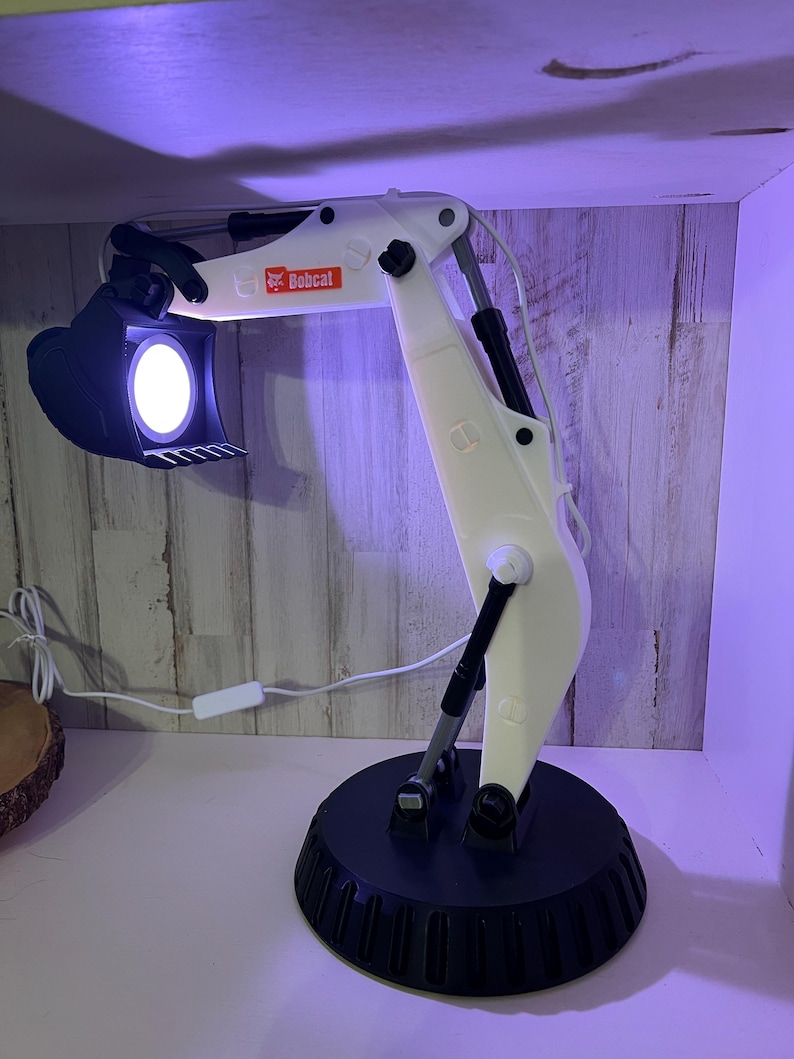 May include: White Bobcat excavator desk lamp with a black base and a white cord. The lamp is turned on and the light is shining from the bucket.