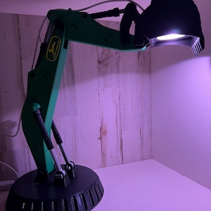 May include: A green John Deere excavator-style desk lamp with a black shade. The lamp is on and the light is shining.
