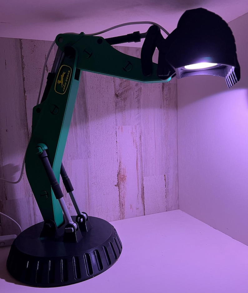 May include: A green John Deere excavator-style desk lamp with a black shade. The lamp is on and the light is shining.