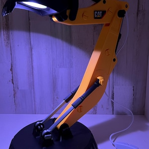 May include: A yellow and black desk lamp shaped like a construction excavator with a black base and a white switch on the cord. The lamp has the word "CAT" printed on the arm.