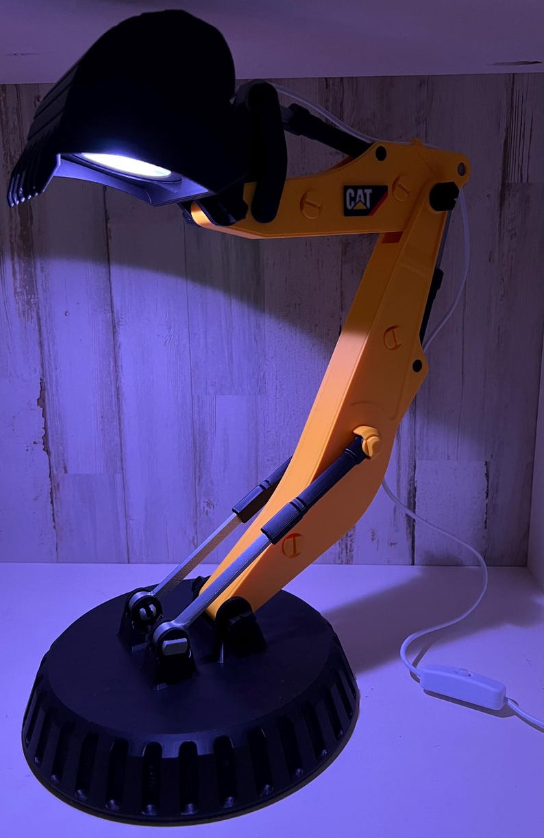 May include: A yellow and black desk lamp shaped like a construction excavator with a black base and a white switch on the cord. The lamp has the word "CAT" printed on the arm.